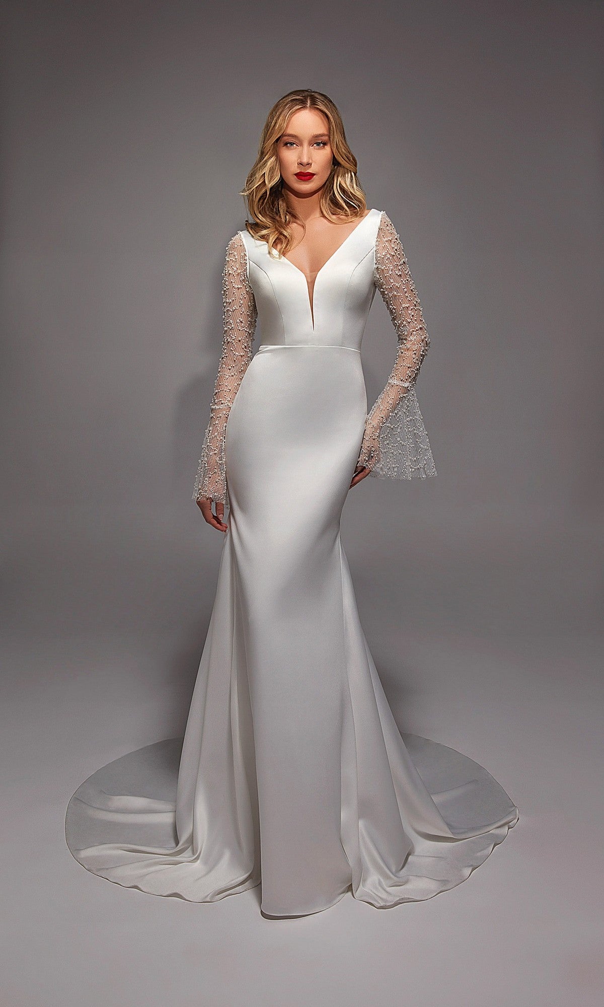 A woman is wearing the Alyce 7137 long white dress in Diamond White, featuring illusion lace sleeves, a deep V neckline, and a long train, standing against a plain gray background.